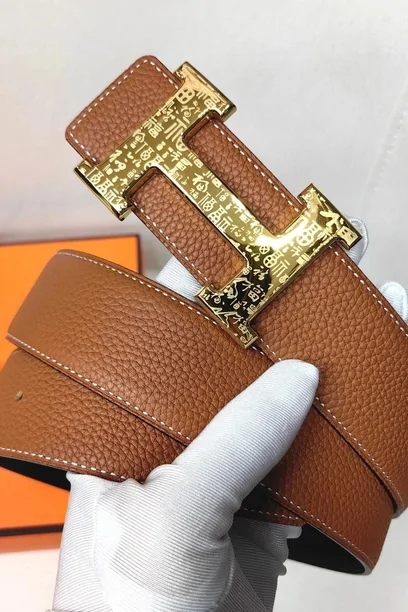 A tan leather belt with a large gold buckle, held up in a gloved hand against an orange backdrop