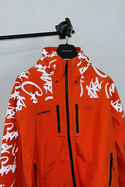 An orange hooded jacket printed with white flame lettering down one sleeve, hanging on a rail
