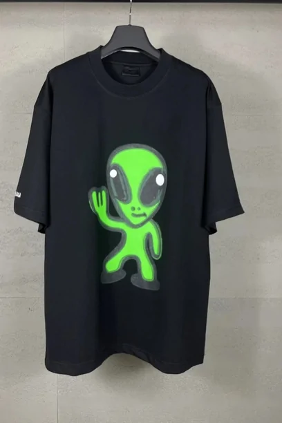 A black tee printed with a large green cartoon alien across the chest, hung against a pale wall